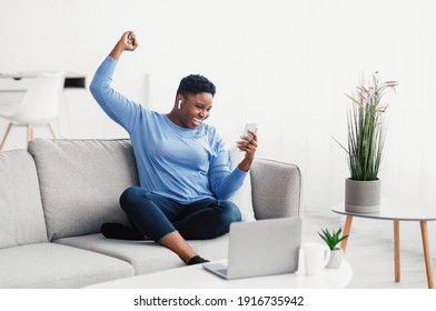 Yes. Excited Overjoyed African American Woman Using Smartphone Gadget, Shaking Clenched Fist, Screaming Yes, Celebrating Victory Or Win, Wearing Wireless Earbuds, Sitting On The Sofa At Home