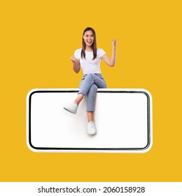 Yes. Excited Woman Sitting On Big Smartphone With Empty White Screen Shaking Clenched Fists, Cheerful Lady Celebrating Win Isolated On Yellow Orange Background, Mock Up Collage, Full Body Length