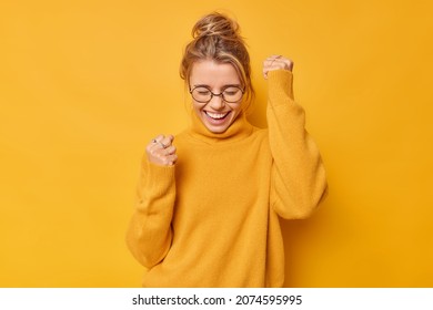 Yes finally success. Positive woman raises arms with clenched fists celebrates something has upbeat mood feels euphoric wears round spectacles and casual jumper poses against bright yellow background