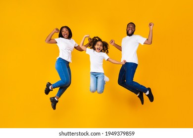 Yes. Full body length of excited young black family of three jumping and shaking clenched fists. Happy African American man, woman and girl celebrating victory together on yellow studio background