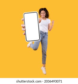 Yes. Full body length portrait of joyful woman jumping up, making winner gesture with clenched fist and showing cellphone with free empty space for mobile app on white screen, orange studio background - Shutterstock ID 2036493071