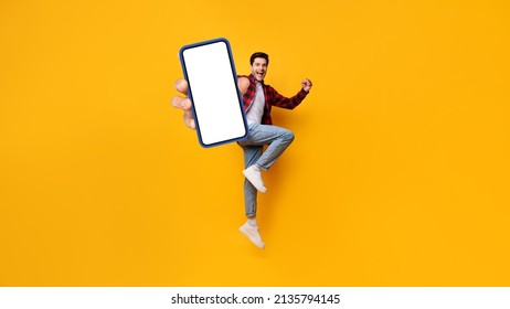 Yes. Full Body Length Portrait Of Joyful Man Jumping Up, Making Winner Gesture With Clenched Fist And Showing Cell Phone With Free Empty Space For Mobile App On White Screen, Yellow Studio Background