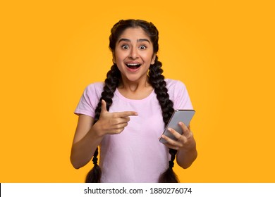 Yes, Great News. Portrait Of Excited Indian Woman Celebrating Win, Using Smartphone And Pointing At Gadget, Smiling Young Lady Having Success, Holding Cellphone, Yellow Studio Background - Powered by Shutterstock