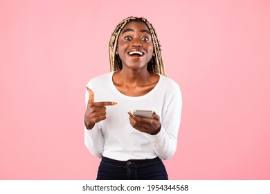 Yes, Great News. Portrait Of Excited Black Woman Celebrating Win, Using Smartphone And Pointing At Gadget, Smiling Young Lady Having Success, Holding Cellphone, Pink Color Studio Background