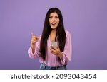 Yes, great news. Surprised armenian woman celebrating win, using smartphone and pointing at gadget, holding cellphone and smiling at camera over purple studio background