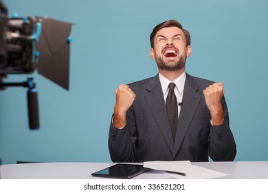 Yes. Handsome Young Tv Newscaster Is Expressing His Happiness. He Is Keeping His Fists And Laughing. The Man Is Sitting At The Table In Front Of The Camera