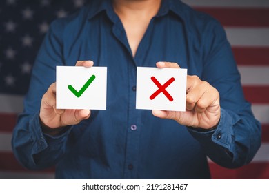 Yes Or No. Close-up Of Hands Holding Two White Papers With A Green Checkmark And Red Cross While Standing On The American Flag Background. True And False Symbols Accept Rejected For Evaluation