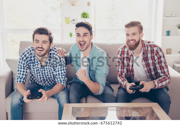 Yes! Our team is winning! Competition of guys\
playing car race. Three excited friends are playing games indoors,\
sitting on cozy beige sofa and enjoying themselves. They have great\
and fun time