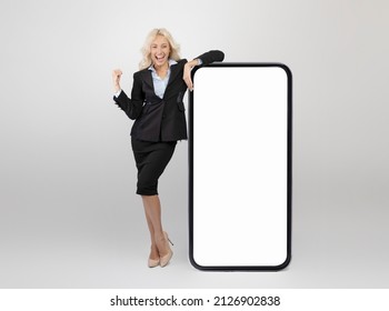 Yes. Overjoyed businesswoman leaning on huge smartphone with empty screen shaking clenched fist, celebrating win, standing over light grey background, mockup collage