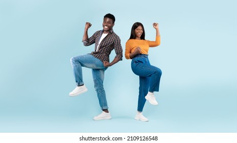 Yes. Overjoyed young black man and woman cheering, jumping and shaking clenched fists. Happy couple celebrating win posing standing isolated over blue studio background wall, banner panorama