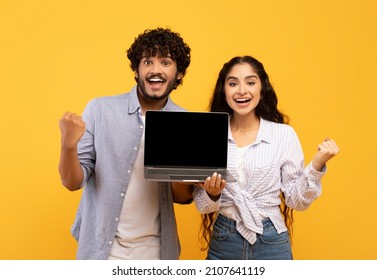 Yes. Portrait Of Happy Indian Couple Holding Laptop With Blank Screen And Shaking Clenched Fists, Showing Computer With Empty Free Space For Mockup, Yellow Studio Background