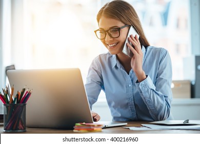 Yes I see your email! Cheerful young beautiful woman in glasses talking on mobile phone and using laptop with smile while sitting at her working place