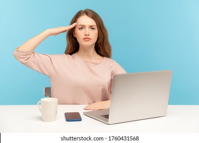 Yes sir! Obedient responsible young woman employee sitting at workplace with laptop and giving salute, listening to boss order, corporate discipline. indoor studio shot isolated on blue background