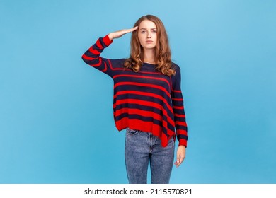 Yes sir Portrait of serious woman wearing striped casual style sweater, standing saluting with hand near head, looking obedient and attentive at camera. Indoor studio shot isolated on blue background