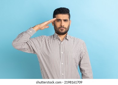 Yes sir. Responsible patriotic businessman saluting with respect as if soldier waiting order from commander, obeying discipline, wearing striped shirt. Indoor studio shot isolated on blue background. - Powered by Shutterstock