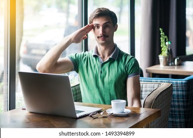 Yes Sir! Young Serious Businessman In Green T-shirt Sitting And Looking At Camera With Salute Gesture And Ready To Complete The Mission. Business Concept. Indoor Shot Near Big Window At Daytime.