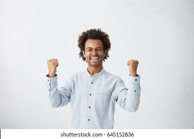 Yes! Waist up studio shot of lucky successful mixed race male student with Afro haircut exclaiming, clenching fists, having winning look after showing the best results on final exams at college
