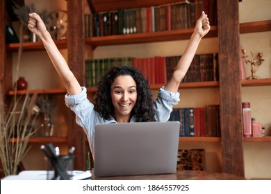 Yes, I'm A Winner. Excited emotional ecstatic lady celebrating success, victory or great news sitting at table, using personal computer, screaming yeah and raising hands up. Smiling happy woman in joy