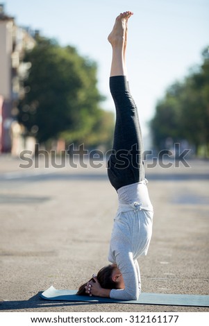 Yoga in the city: beautiful young sporty woman working out on the road on summer day, doing supported headstand posture, salamba sirshasana, full length, profile view