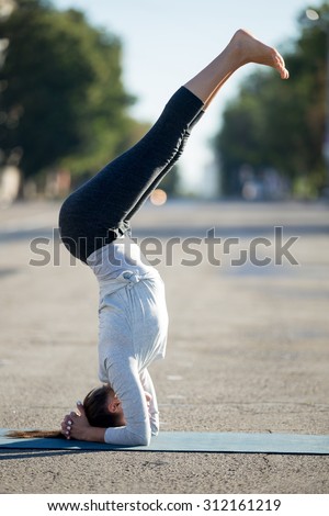 Yoga in the city: beautiful young sporty woman doing supported headstand posture, variation of salamba sirsasana in the street on summer day, full length, side view