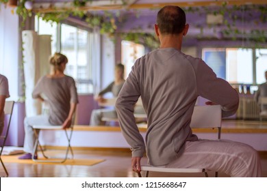 Yoga class, Half Lord of the Fishes Pose (Ardha Matsyendrasana) done by female and male practitioner sat on a chair with mirror in the backgroung.