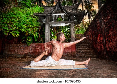 Yoga Eka Pada Raja Kapotasana By Fit Man In White Trousers Near Stone Temple In Tropical Forest