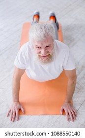 Yoga Enthusiast. The Top View Of A Handsome Elderly Man Smiling Pleasantly While Standing In Upward-facing Dog Pose On A Yoga Mat