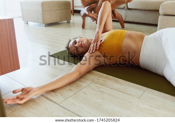 Yoga Exercises. Woman In\
Reclining Spinal Twist Position. Female In Fashion Sportswear\
Practicing Supta Matsyendrasana At Home. Sport Routine For Active\
Lifestyle.