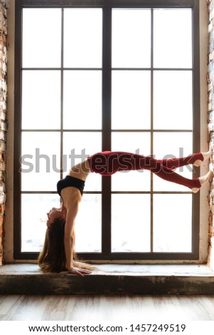 Yoga girl in a black top and red leggings stands on her hands on the windowsill of a large window