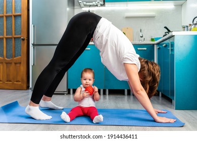 Yoga at home. A young mother is doing Adho Mukha Shvanasana, her baby is sitting on the mat. Side view. The concept of fitness with kids. - Shutterstock ID 1914436411