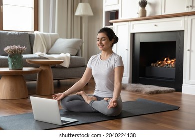 Yoga at home. Young sporty indian female practice sukhasana position meditate on carpet before modern laptop computer at luxury living room. Fit mixed race woman do gymnastic exercises indoor using pc
