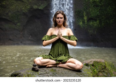 Yoga lotus pose. Young Caucasian woman sitting on the stone, meditating, practicing yoga, pranayama at waterfall. Hands in namaste mudra. Closed eyes. Yoga retreat. Tibumana waterfall, Bali, Indonesia