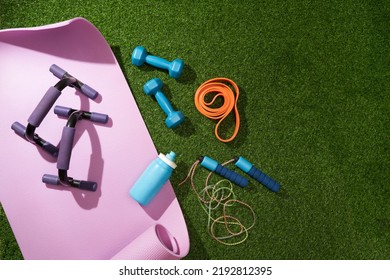 Yoga Mat With Dumbbells, Jump Rope, Headphones, Water Bottle And Other Sport Gear On Green Grass.