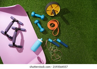 Yoga Mat With Dumbbells, Jump Rope, Headphones, Water Bottle And Other Sport Gear On Green Grass.