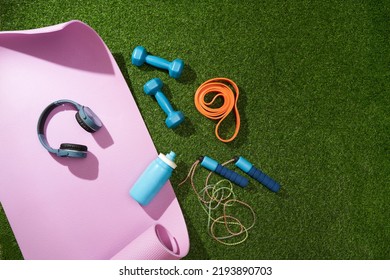 Yoga Mat With Dumbbells, Jump Rope, Headphones, Water Bottle And Other Sport Gear On Green Grass.