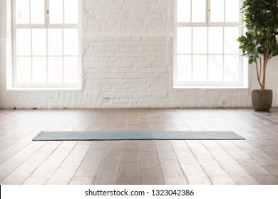 Yoga mat on natural wooden floor in empty room in fitness center, comfortable space for doing sport exercises, big windows and white brick walls, modern yoga class room with nobody