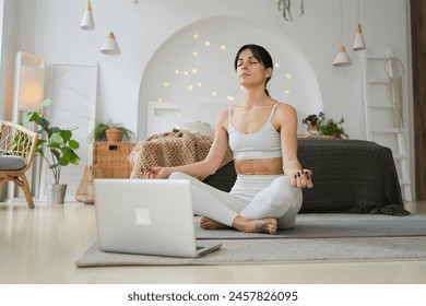 Yoga mindfulness meditation online. Woman practicing yoga with online lessons in laptop at home. Woman sitting in lotus pose meditating relaxing indoor. Girl doing breathing practice. Yoga at home - Powered by Shutterstock