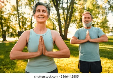 Yoga on retirement. Front view of mature man and woman dressed in black and blue outfits keeping hands in namaste gesture and meditating outdoors. Smiling couple closing eyes and doing yoga exercises. - Powered by Shutterstock