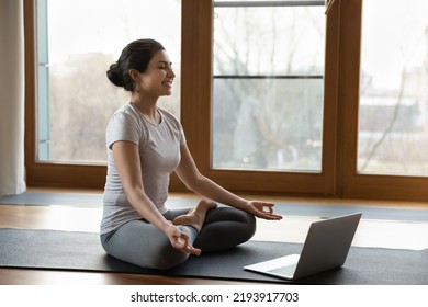 Yoga online. Happy young Indian female yogi do exercises before laptop screen at home or in gymnastic studio. Smiling millennial woman coach sit in lotus position practice sukhasana give web - Powered by Shutterstock