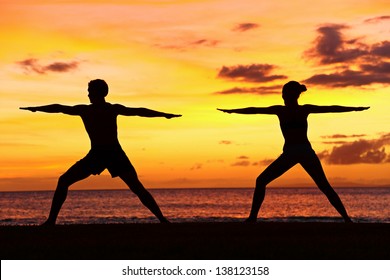 Yoga People Training And Meditating In Warrior Pose Outside By Beach At Sunrise Or Sunset. Woman And Man Yoga Exercising Training In Serene Ocean Landscape. Silhouette Of Couple Against Sun.