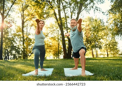 Yoga practice outdoors. Front view of encouraged male and female adults staying in warrior pose on rugs. Focused family partners dressed in activewear outstretching arms and looking straight. - Powered by Shutterstock