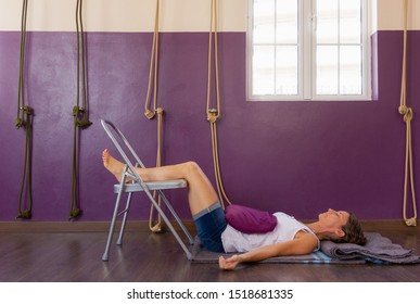 Yoga student rests on floor with legs up on chair at studio. Female yogi with sand bag on stomach at therapeutic class with ropes on purple wall background. Healthy lifestyle, pain, healing concepts