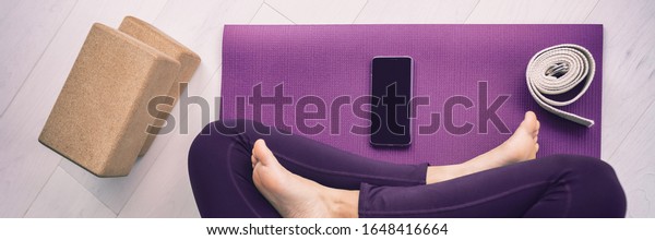 Yoga woman\
using mobile phone panoramic banner top view of cellphone on\
exercise mat and blocks, stretching\
strap.