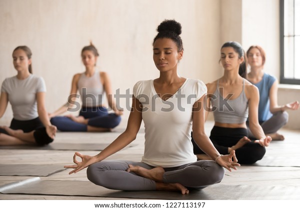 Йоги черная женщина и разнообразная группа молодых спортивных людей, практикующих йогу, делая Padmasana упражнения, Lotus позы, работая в помещении, студентки обучение в клубе. Концепция хорошего самочувствия