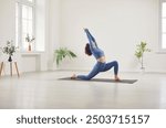 Yogi sporty attractive woman practicing yoga, doing Horse rider exercise, anjaneyasana pose, working out alone, wearing elegant sportswear in white yoga studio, among indoor green houseplants