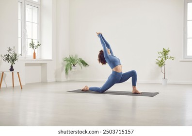 Yogi sporty attractive woman practicing yoga, doing Horse rider exercise, anjaneyasana pose, working out alone, wearing elegant sportswear in white yoga studio, among indoor green houseplants