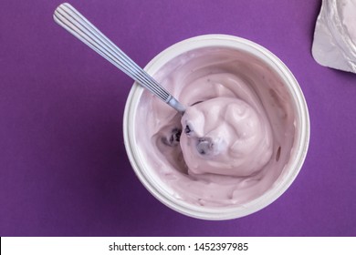 Yogurt cup with blue berry yoghurt, spoon and foil lid isolated on purple background - top view photograph