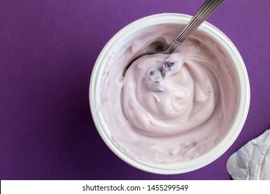 Yogurt cup with blue berry yoghurt, spoon and foil lid isolated on purple background with text space - top view photograph of creamy fruit yoghurt