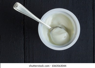 Yogurt Cup With Natural Greek Yoghurt In Plastic Cup With Silver Spoon Closeup On Rustic Black Table Background - Top View Photo