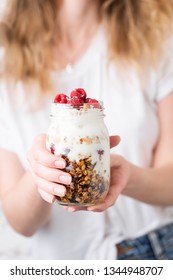 Yogurt With Granola And Berries In Jar In Female Hands. Blonde Woman In White Shirt Holding Jar Of Yogurt Parfait With Granola And Berries. Healthy Eating, Lifestyle, Trendy Vegetarian Food Concept
